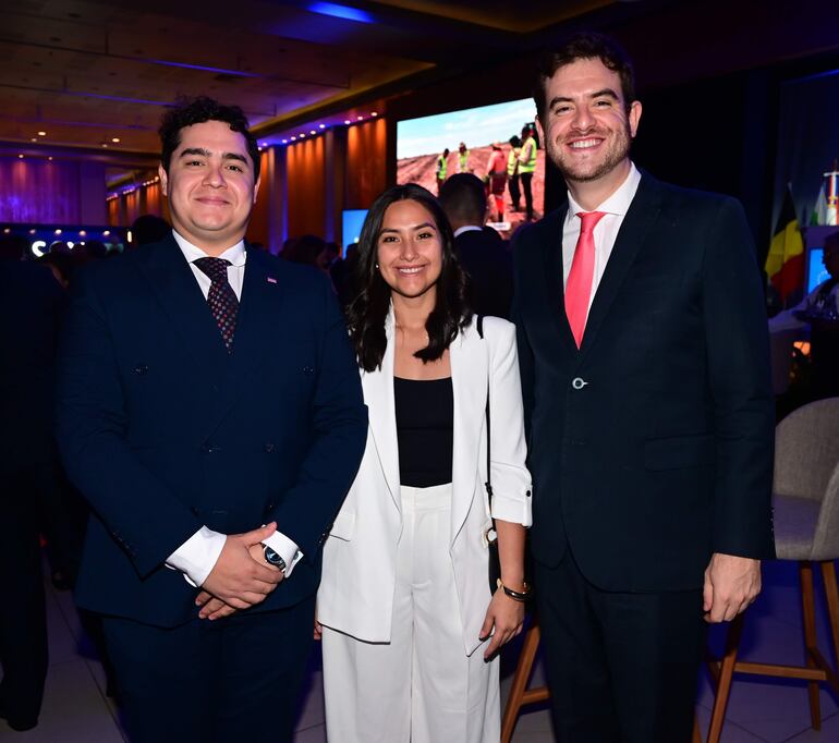 Hombre en traje oscuro y corbata, mujer con conjunto blanco y blusa negra, y otro hombre con traje oscuro y corbata roja, todos sonriendo.
