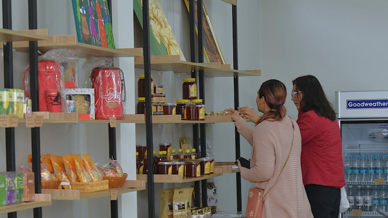 Dos mujeres, una con suéter claro y otra con gafas, observan productos en estanterías de madera en un ambiente iluminado.