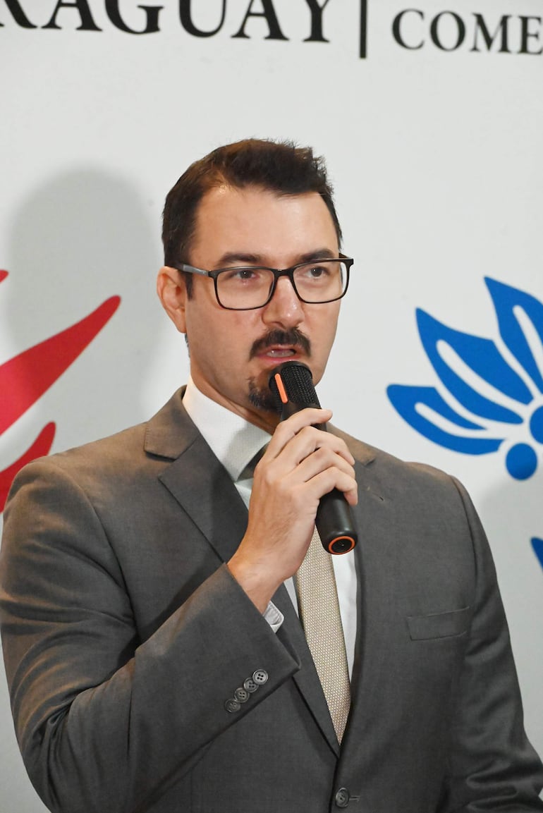 Hombre en traje gris, con micrófono, habla en evento formal. Fondo con logotipo, ambiente interior y público ausente.