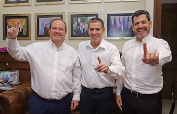 Camilo Pérez, Raúl Latorre y Horacio Cartes con camisas blancas y sonrisa, levantando el dedo índice en un fondo decorado formalmente.