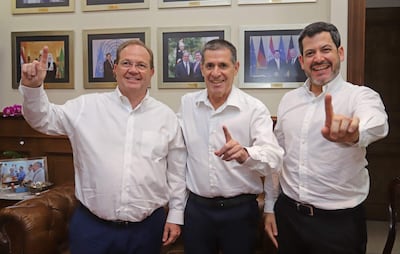 Camilo Pérez, Raúl Latorre y Horacio Cartes con camisas blancas y sonrisa, levantando el dedo índice en un fondo decorado formalmente.