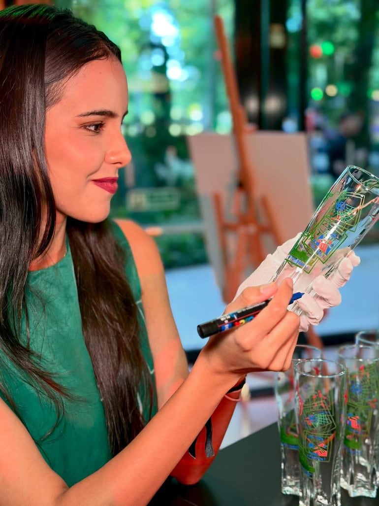 Mujer de cabello oscuro y blusa verde, firmando vaso con marcador, guante en mano izquierda, ambiente de evento interior.
