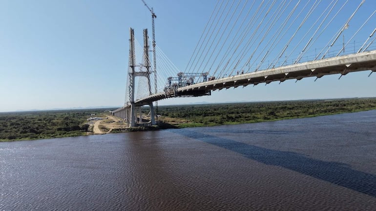 Puente en construcción sobre un río con maquinaria visible, rodeado de un entorno natural y bajo un cielo despejado.