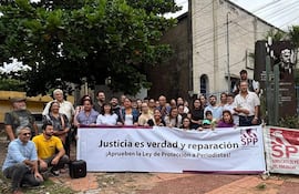 Grupo de personas sosteniendo un banner, algunos con expresiones de tristeza y alegría en un acto de protesta en un espacio urbano.