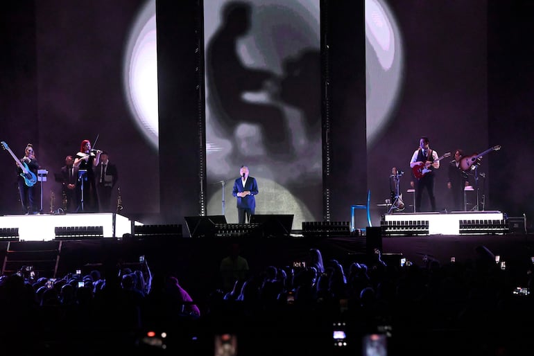 Ricardo Montaner en traje oscuro y camisa blanca, rodeado de músicos en un vibrante concierto con luces brillantes.