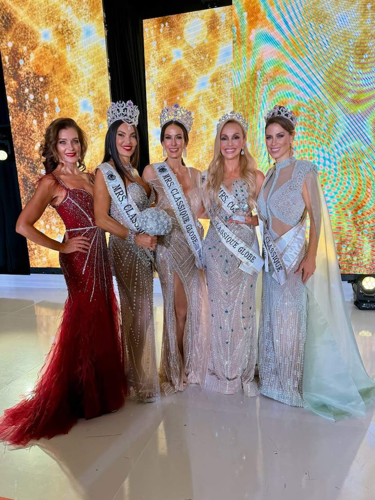 Cinco mujeres sonrientes en un certamen de belleza, vestidas elegantemente, con coronas y bandas, posando en un fondo festivo.