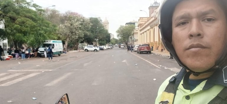 Hombre con casco y expresión seria en la calle, rodeado de grupos de personas y un vehículo de emergencias al fondo.