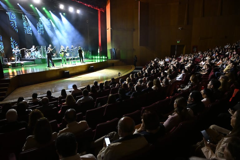 Músicos de Los Nocheros en el escenario, con luces coloridas y una audiencia de 200 personas en butacas rojas.