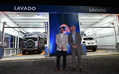 Dos hombres formales, uno con saco claro y otro con saco gris, junto a instalaciones de lavado de autos modernas.