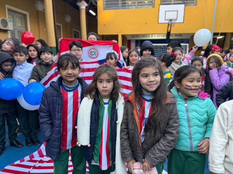 Niñas sonrientes en camisetas de Paraguay y abrigos de colores, sosteniendo una bandera paraguaya y globos en un ambiente festivo.