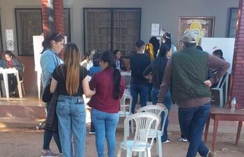 Mujeres conversando en un centro de votación, con un hombre de espaldas observando. Ambiente exterior, mesas de votación visible.