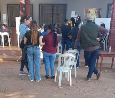 Mujeres conversando en un centro de votación, con un hombre de espaldas observando. Ambiente exterior, mesas de votación visible.