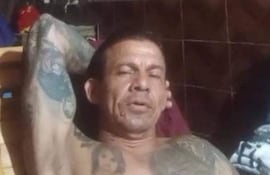 Hombre relajado y sentado con torso descubierto y tatuajes visibles en ambiente interior de vivienda común.