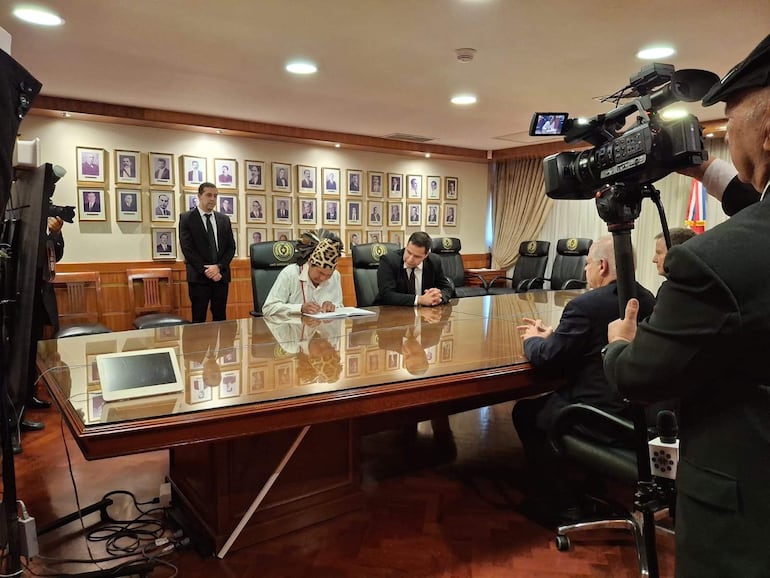 Hombre Ayoreo en tocado de plumas firma documentos, rodeado de hombres en traje en sala formal con retratos en la pared.