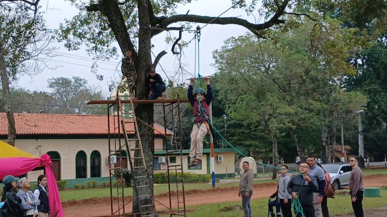 Hombre con casco y arnés se lanza en tirolesa mientras otros observan, en un entorno natural con vegetación y un edificio de fondo.