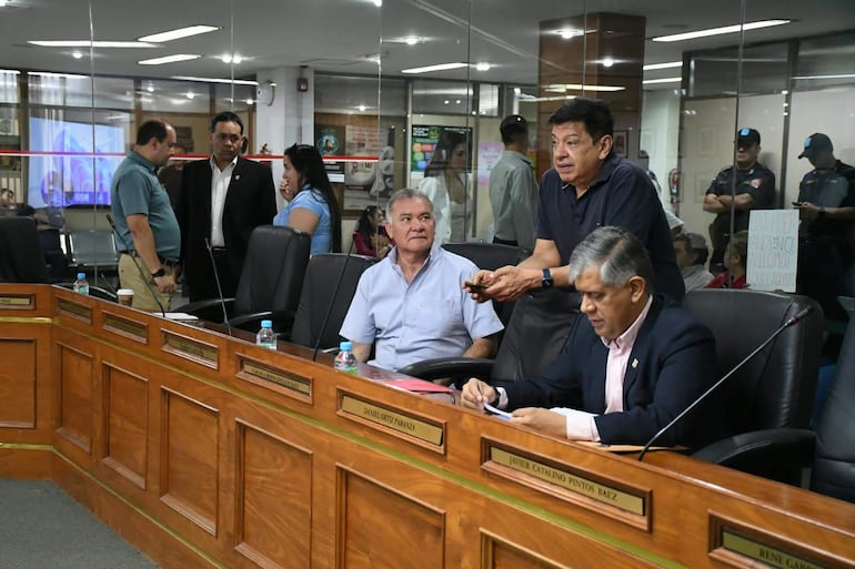 Augusto Wagner de pie sosteniendo un teléfono, Felix Ayala sentado mirando documentos, ambiente interior con varias personas.