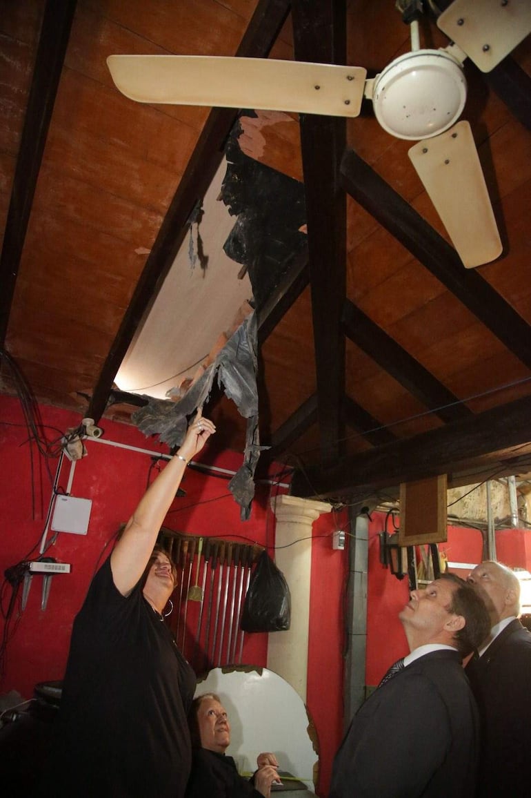 Una mujer de pie señala el techo dañado, acompañada de tres hombres, en un ambiente con paredes rojas y decoración simple.