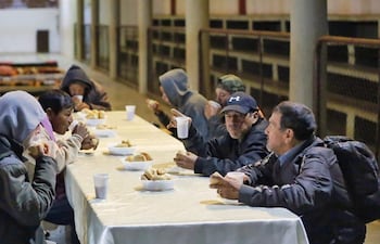 Diez personas consumiendo comida en un comedor, vistiendo ropa abrigada en ambiente tenue y de comunidad.