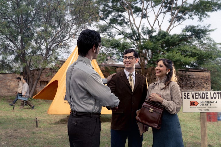 Tres personas: hombre de camisa clara conversando, hombre con gafas sonriendo y mujer en blusa de puntos, junto a carpa naranja y cartel.