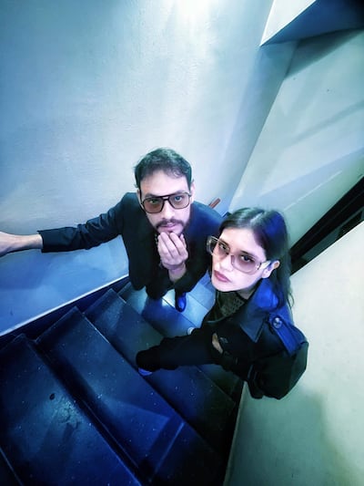Dos integrantes del dúo Sandía, Wilson Santos con traje oscuro y gafas, y Belén Pinto mujer en abrigo negro, posan en una escalera con expresión neutral.