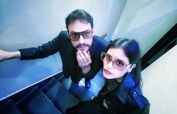 Dos integrantes del dúo Sandía, Wilson Santos con traje oscuro y gafas, y Belén Pinto mujer en abrigo negro, posan en una escalera con expresión neutral.