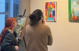 Dos mujeres observan pinturas coloridas en la galería, una con abrigo oscuro y la otra con suéter claro, en un ambiente de arte.