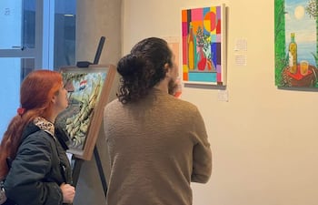 Dos mujeres observan pinturas coloridas en la galería, una con abrigo oscuro y la otra con suéter claro, en un ambiente de arte.