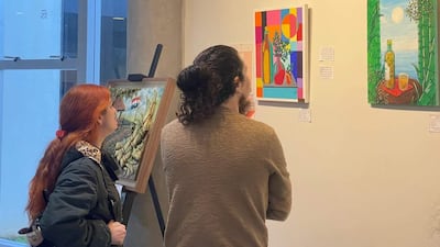Dos mujeres observan pinturas coloridas en la galería, una con abrigo oscuro y la otra con suéter claro, en un ambiente de arte.