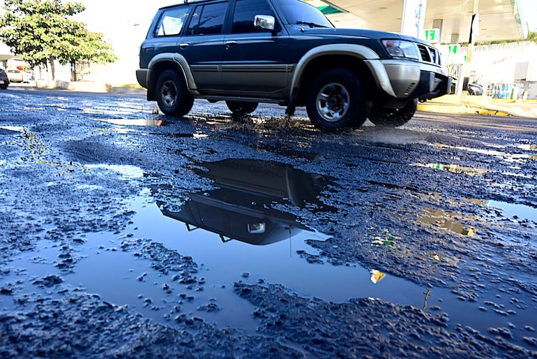 Automóvil azul oscuro en movimiento, dispersando agua de charcos en calle pavimentada y dañada, sin personas visibles.
