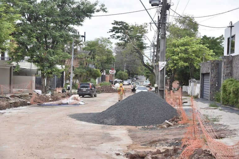 Trabajador en chaleco de seguridad y casco amarillo supervisa obra en calle, con montículo de grava y vehículos estacionados al fondo.