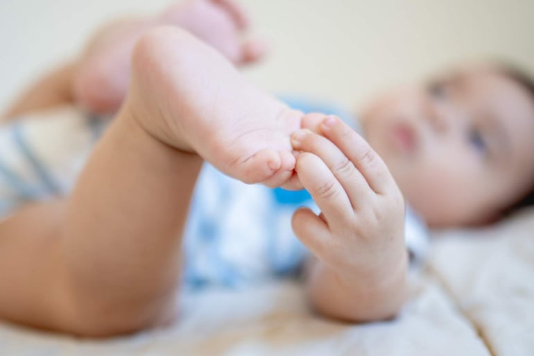 Niño pequeño en superficie clara, abrazando su pie con una mano, con gesto tranquilo.