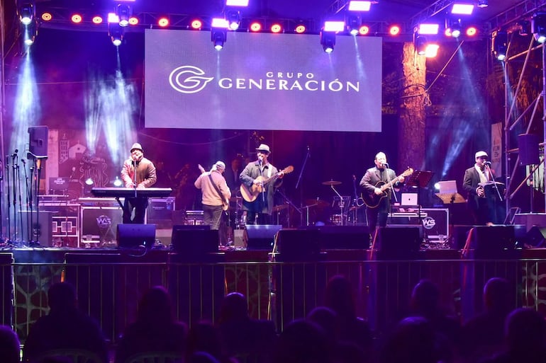 Músicos del grupo Generación en el escenario, tocando guitarra y teclado con iluminación colorida y público atento.