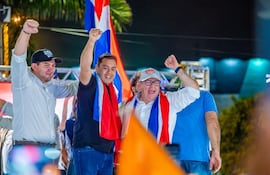 Tres hombres levantan el puño en celebración, uno viste camiseta blanca y gorra, otro camiseta oscura con bandera paraguaya.