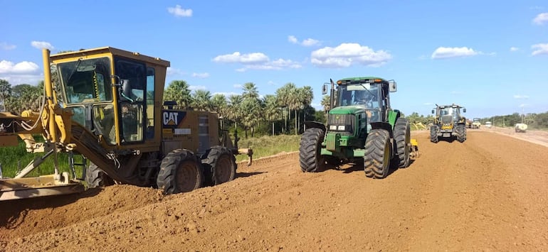 Máquinas pesadas en obra de pavimentación, un tractor verde y un bulldozer operando en terreno de tierra bajo un cielo azul.