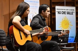 Hombre y mujer con guitarras vestidos de negro, interpretando música en un escenario formal, con banner de 'HA CHE VALLE' detrás.
