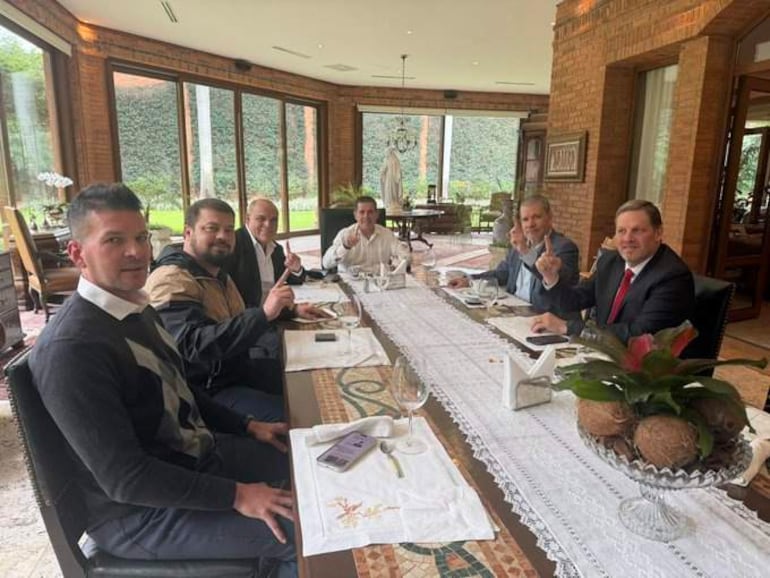 Horacio Cartes y Bachi Núñez rodeados de aliados, todos vestidos formalmente, intercambiando gestos de acuerdo en una mesa bien decorada.