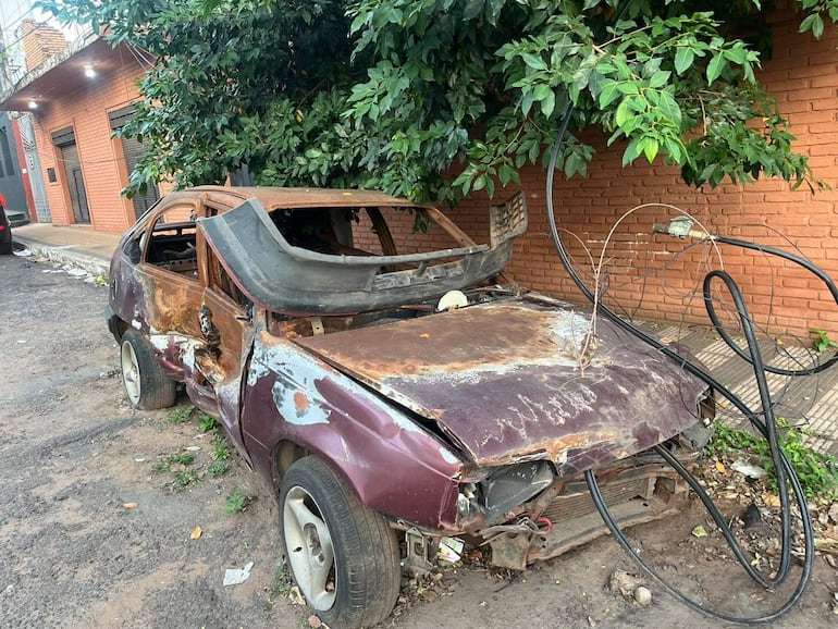 Vehículo dañado de color púrpura, con carrocería oxidada, rodeado de plantas en ambiente urbano descuidado.