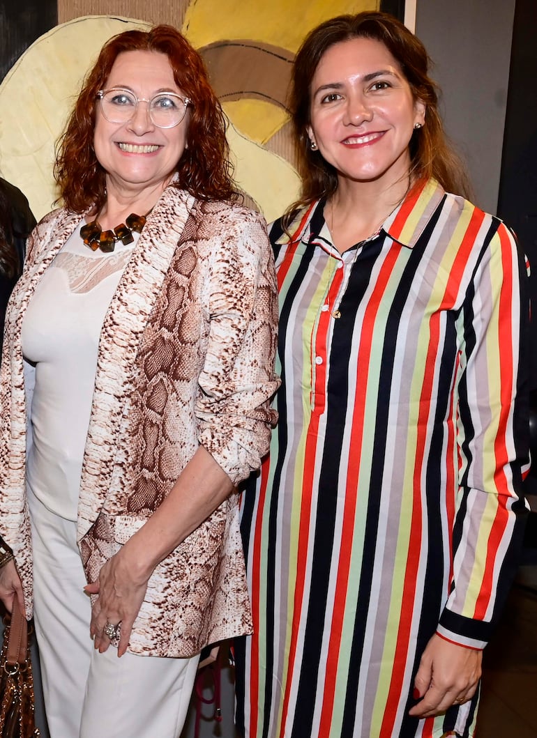 Dos mujeres sonrientes: una con chaqueta de serpiente y blusa blanca, y la otra en camisa de rayas, posando en un ambiente decorado con arte abstracto.