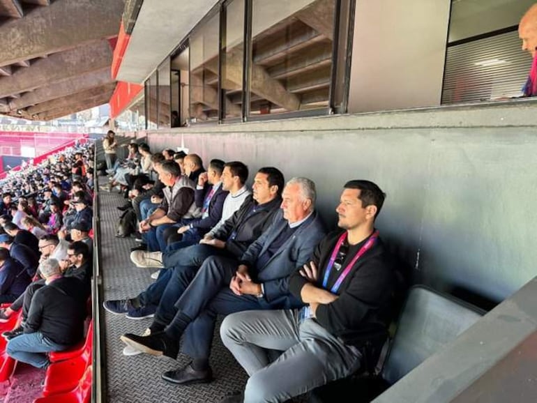 Robert Harrison y otros espectadores, todos en ropa casual, observan atentamente un evento en un estadio.