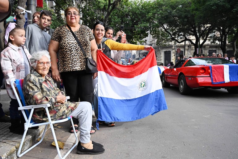 Grupo de seis personas, dos con bandera de Paraguay, una mujer mayor con abrigo camuflado y expresión sonriente, en un ambiente festivo.