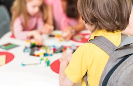 Imagen de un niño de espaldas con su mochila sentado frente a una niña y la profesora en el pre escolar.