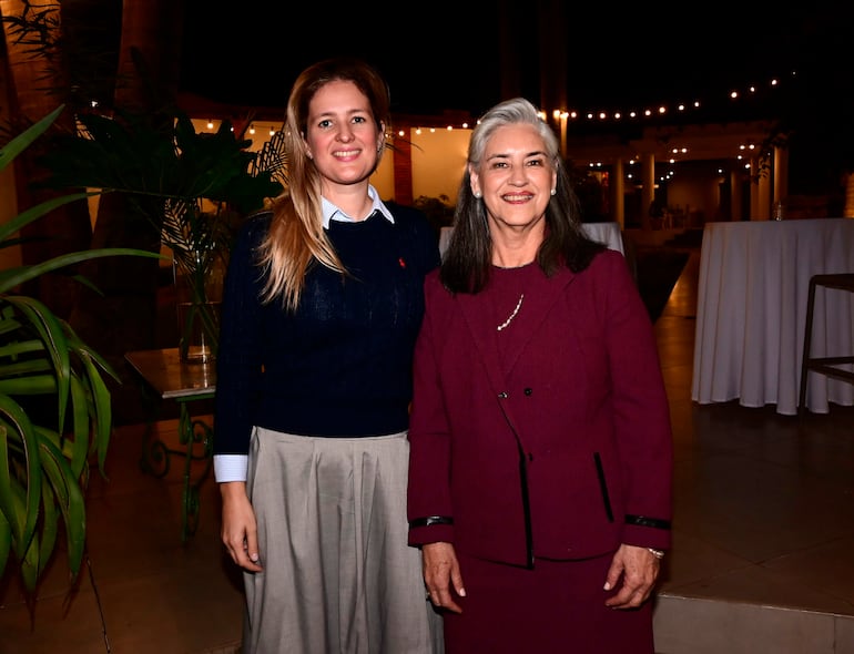 Cristina Goralewski sonríe junto a otra mujer, vestida elegantemente en un evento social con luces cálidas y decoración festiva.