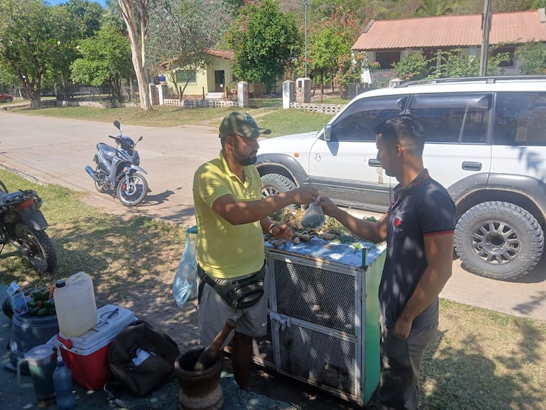 Hombre en camisa amarilla y gorra conversa con cliente, a su lado un puesto con productos frescos en entorno rural.