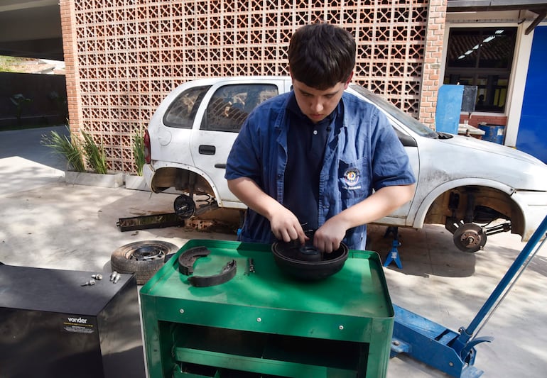 Joven concentrado en taller mecánico, manipulando pieza de freno sobre mesa verde, con auto blanco de fondo.