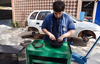 Joven concentrado en taller mecánico, manipulando pieza de freno sobre mesa verde, con auto blanco de fondo.