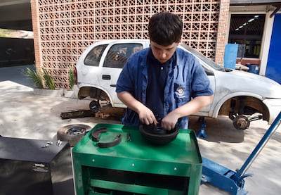 Joven concentrado en taller mecánico, manipulando pieza de freno sobre mesa verde, con auto blanco de fondo.
