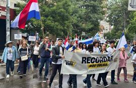 Una mujer mayor sonriente con blusa rosa sostiene junto a un hombre pancarta que dice 'RECORDAR NOS OBLIGA' en una marcha pacífica.