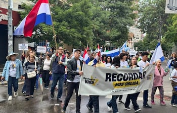 Una mujer mayor sonriente con blusa rosa sostiene junto a un hombre pancarta que dice 'RECORDAR NOS OBLIGA' en una marcha pacífica.