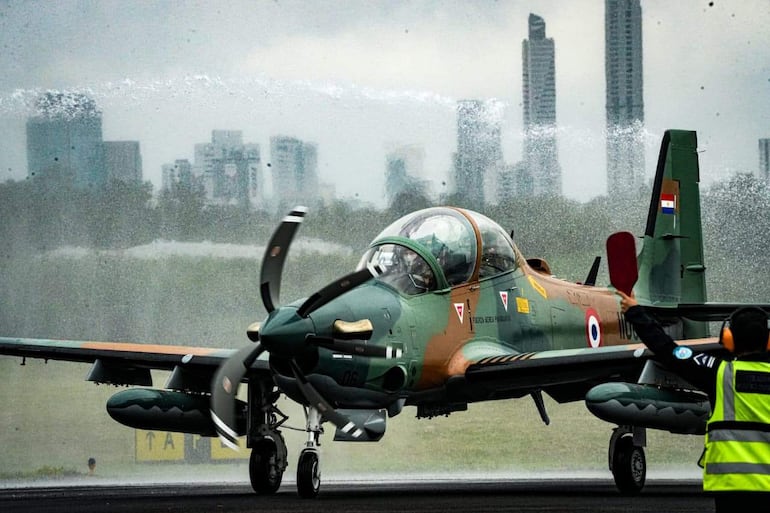 Un operador de tierra con chaleco reflectante y auriculares dirige un Super Tucano en un cielo nublado, mientras edificios urbanos se ven al fondo.