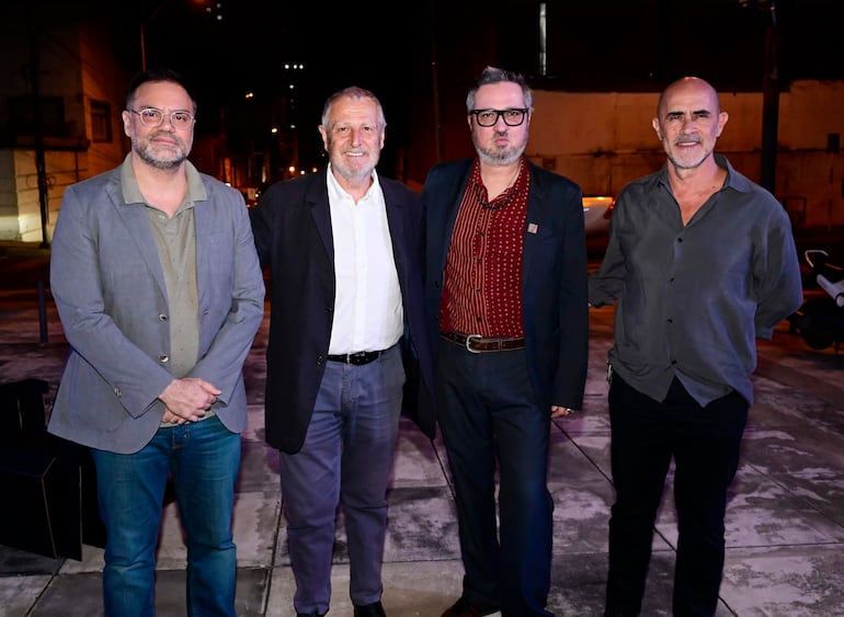 Martín Nasta Bittar sonríe junto a tres hombres en chaquetas, en ambiente nocturno con luz artificial.
